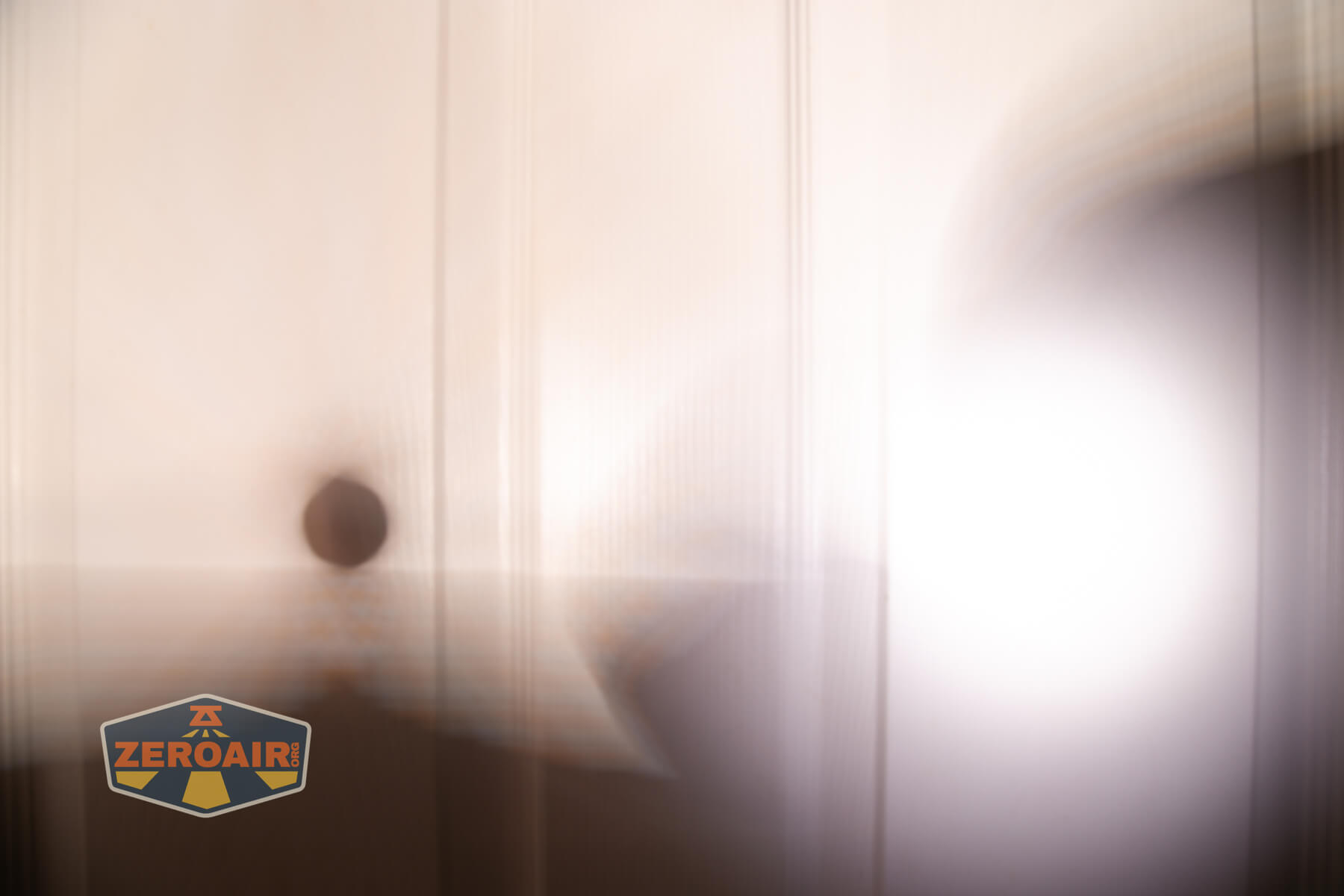 A blurred image showing a white background with vertical lines, a round dark spot, and a bright white light. The lower left corner has a logo with the text ZEROAIR.